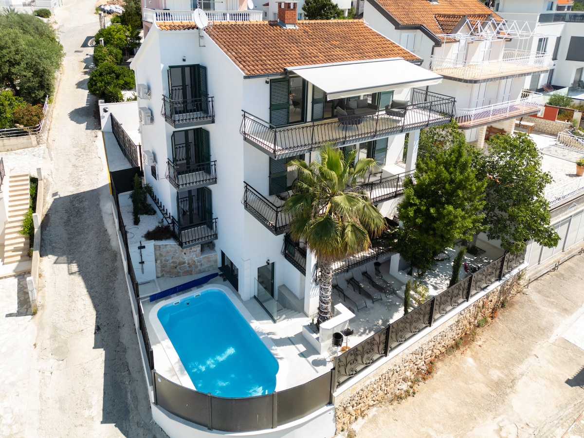 A modern three-story building features a well-maintained swimming pool surrounded by a patio area with sun loungers. Lush green palm trees add a touch of nature, while various balconies provide outdoor seating, enhancing the inviting surroundings.