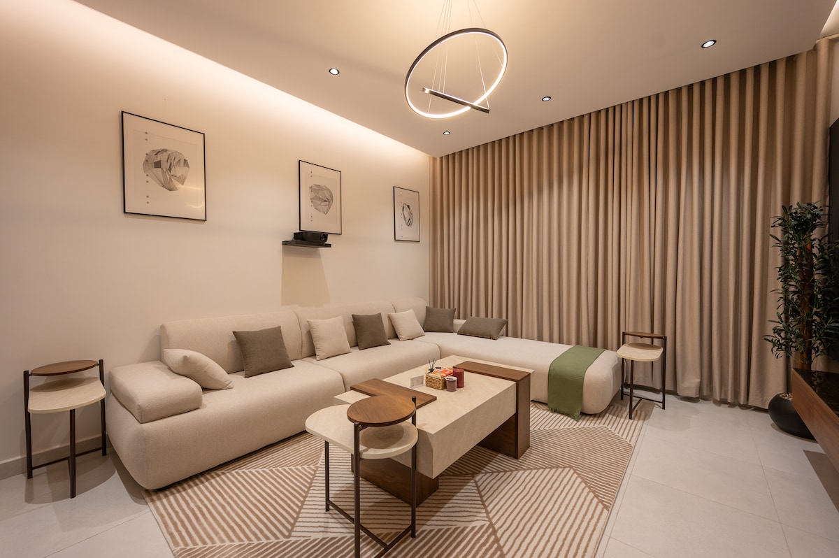 A spacious living area features a large sectional sofa complemented by neutral-toned cushions. A geometric area rug anchors the space, while a wooden coffee table adds warmth. Wall art is displayed above, and soft curtains frame the windows, creating an inviting ambiance.