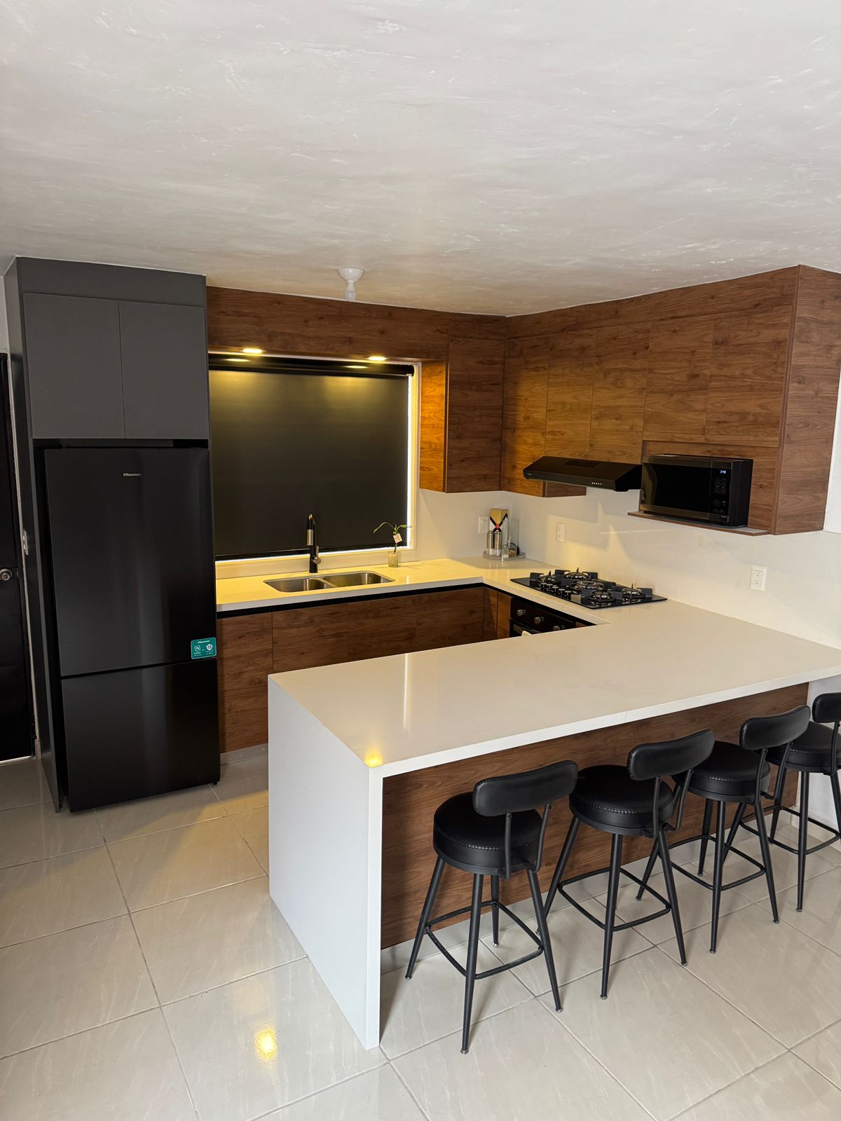 A modern kitchen is seen, featuring a sleek white countertop paired with warm wood cabinetry. Black bar stools are arranged at the island, while integrated appliances include a microwave and stove. The under-sink area is well-organized, with ample natural light coming from the window.