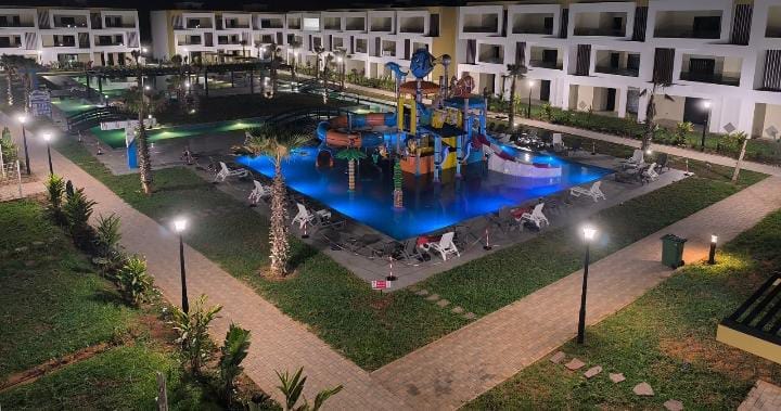 A large outdoor area features a swimming pool with colorful water play structures and illuminated by soft lighting. Sun loungers line the poolside, surrounded by landscaped greenery. The spacious layout is complemented by walking paths and palm trees, creating a family-friendly environment.