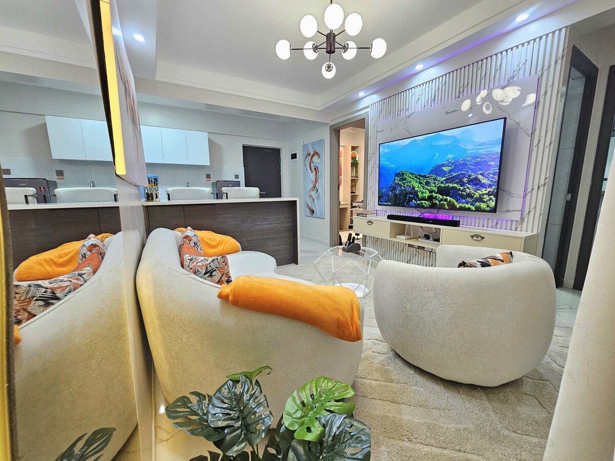 A contemporary living area features white, curved seating adorned with colorful cushions. A large Smart TV is mounted on the wall, displaying vibrant scenery. A neutral-toned area rug covers the floor, while indoor plants add a touch of greenery, contributing to a cozy atmosphere.