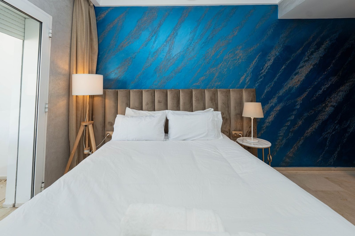 Hoofdslaapkamer met een queensize bed, getextureerde blauwe muur, zachte verlichting en verduisteringsgordijnen. Comfort en stijl gecombineerd, gezellige sfeer en praktische opslag.