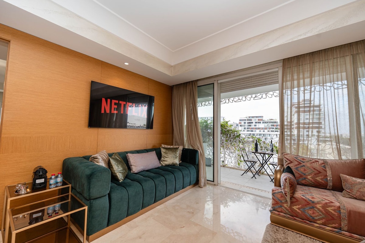 A refined living area features a plush green sofa contrasted with decorative pillows. A wall-mounted Smart TV displays the Netflix logo. Sliding glass doors provide access to a balcony with a small table and chairs. Warm natural light filters through sheer curtains.