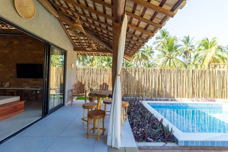 An outdoor patio area features wooden chairs surrounding a round table, situated next to a refreshing pool. Tropical vegetation and palm trees are visible in the background, complemented by a rustic wooden structure with an open roof. Soft natural light enhances the serene atmosphere.
