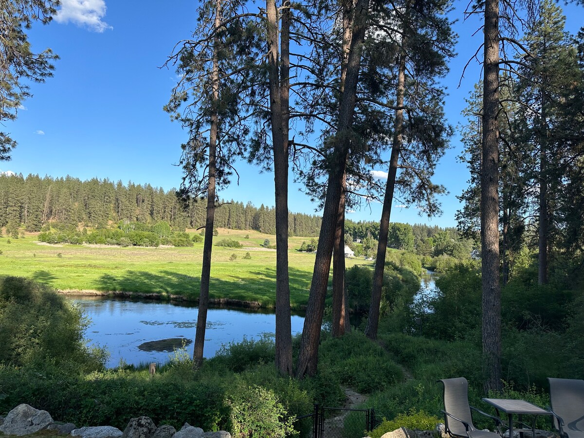 A tranquil view of the Little Spokane River is framed by tall pine trees. Lush greenery and rolling hills are visible in the background, with the river reflecting the clear blue sky. Outdoor seating for relaxation is positioned in the foreground.