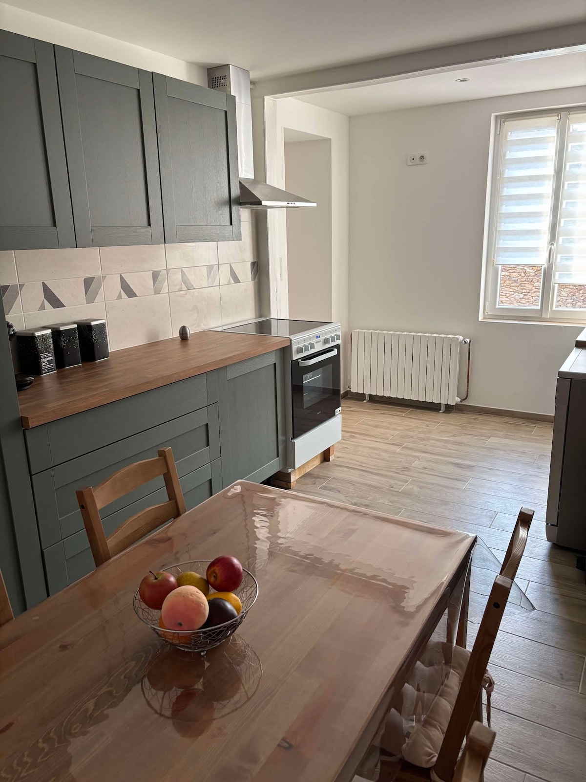 A modern kitchen features green cabinetry and a wooden countertop. A dining table is set with a bowl of apples, while a window allows natural light to fill the space. Appliances include an oven and a radiator along the wall, enhancing functionality.