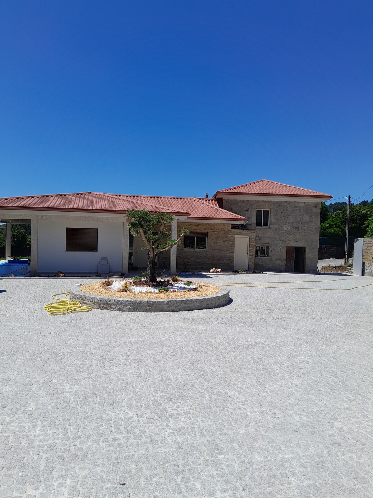 A modern stone building with a red-tiled roof is surrounded by a paved area. A circular garden bed with decorative stones and an olive tree is positioned prominently in the foreground. Two entrances and multiple windows are visible, providing a bright and open appearance.
