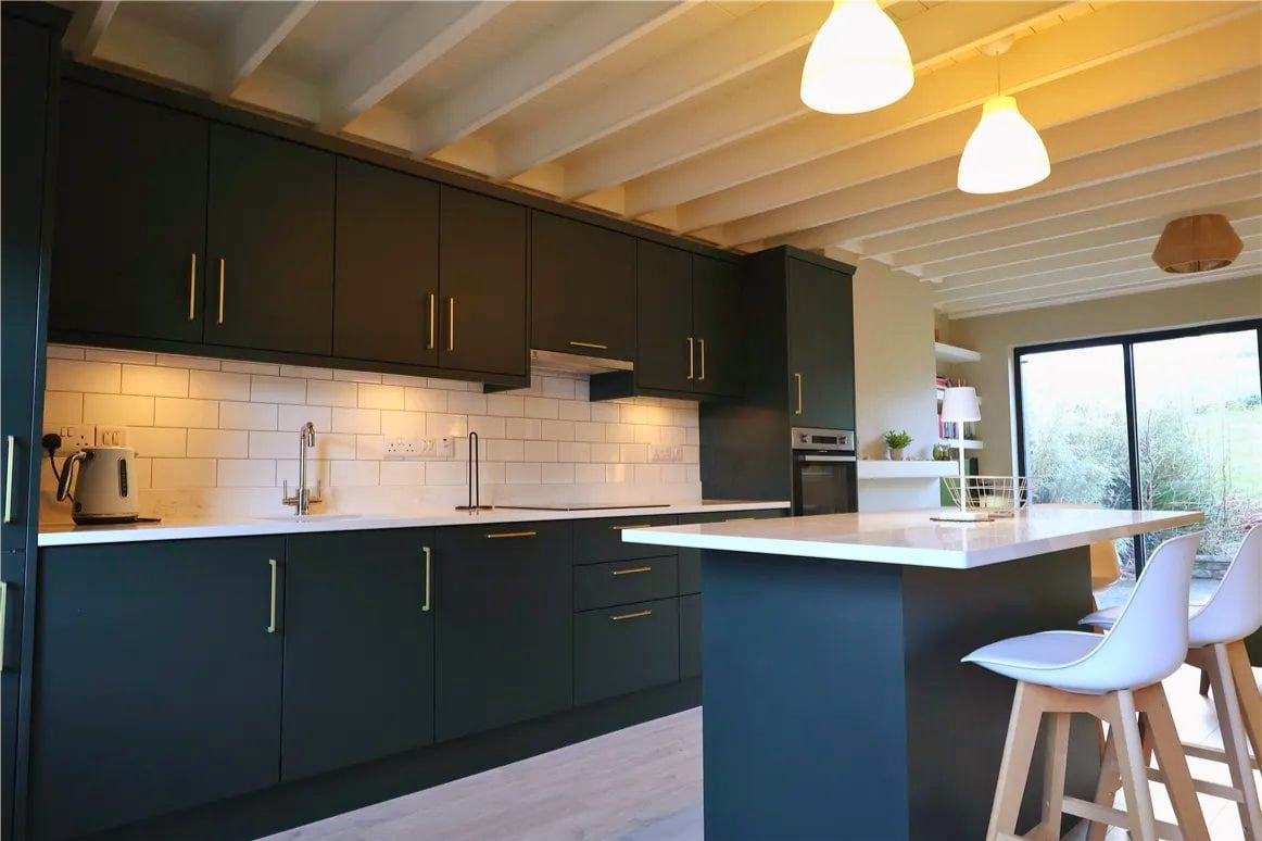 A modern kitchen features dark cabinetry complemented by light countertops. A spacious island provides additional dining space, while pendant lighting hangs from the ceiling. Natural light floods in through large glass doors that lead to an outside terrace, enhancing the kitchen's bright atmosphere.