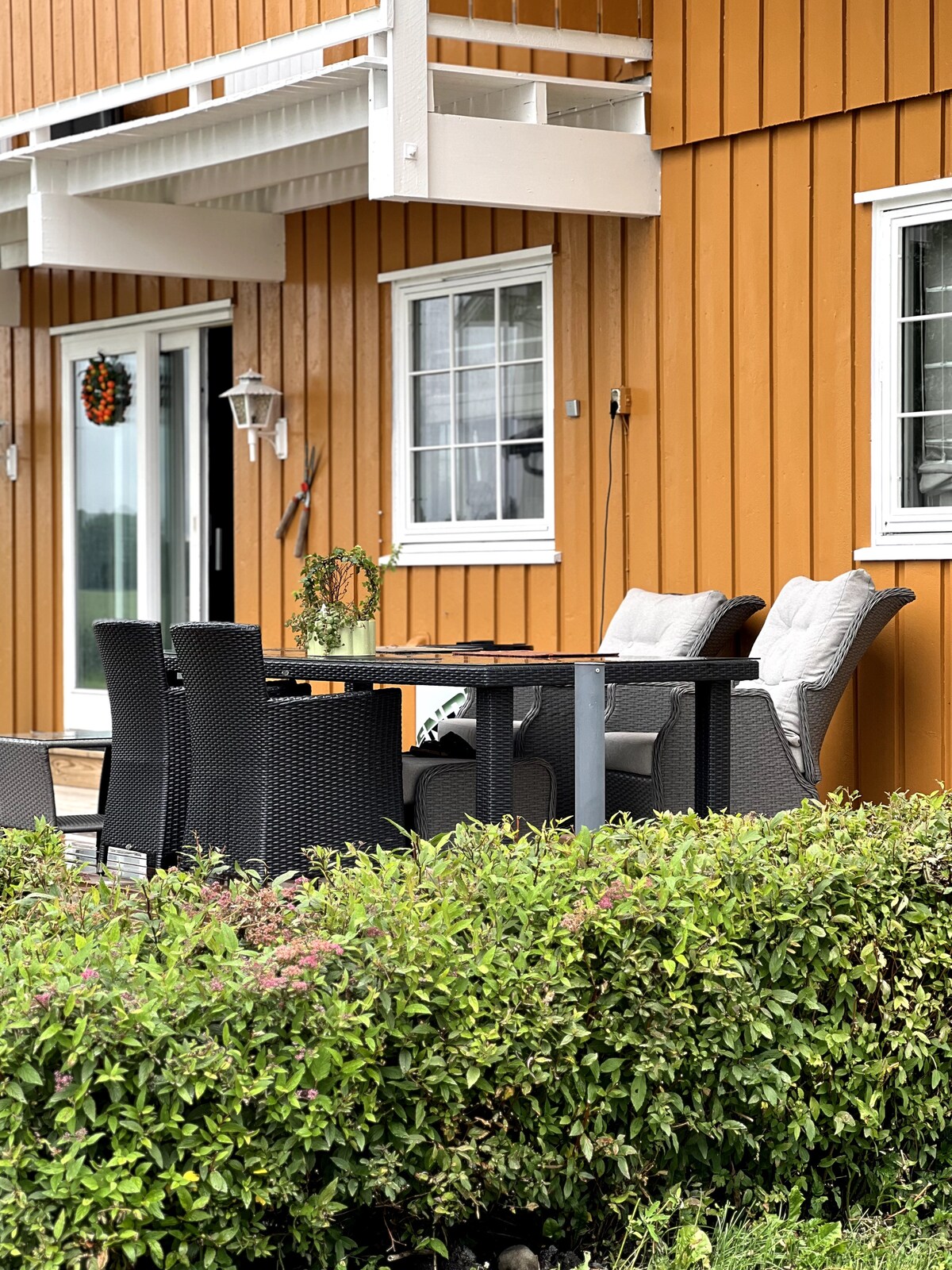 An outdoor seating area features a rectangular table surrounded by four wicker chairs, each adorned with soft cushions. A decorative wreath hangs on the wall, and the vibrant orange exterior of the building contrasts with the lush green foliage nearby.