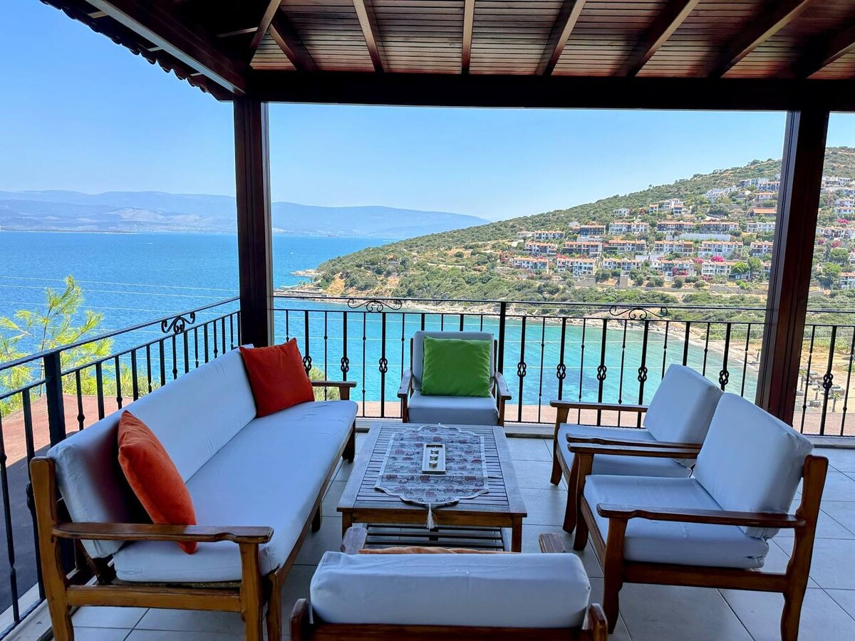 A shaded balcony presents a comfortable seating arrangement with white sofas and colorful cushions. A small table is set at the center. The stunning view includes a clear blue sea and a hillside dotted with vibrant homes, enhancing the outdoor experience.
