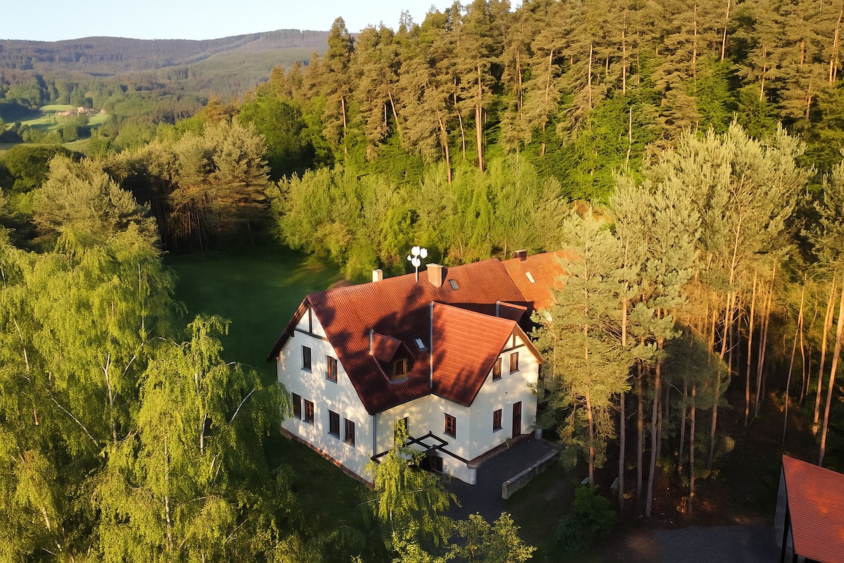 The exterior of a large building is surrounded by lush greenery and tall trees. The structure features a red roof and multiple windows. A spacious lawn is visible in front, complemented by a natural landscape in the background.