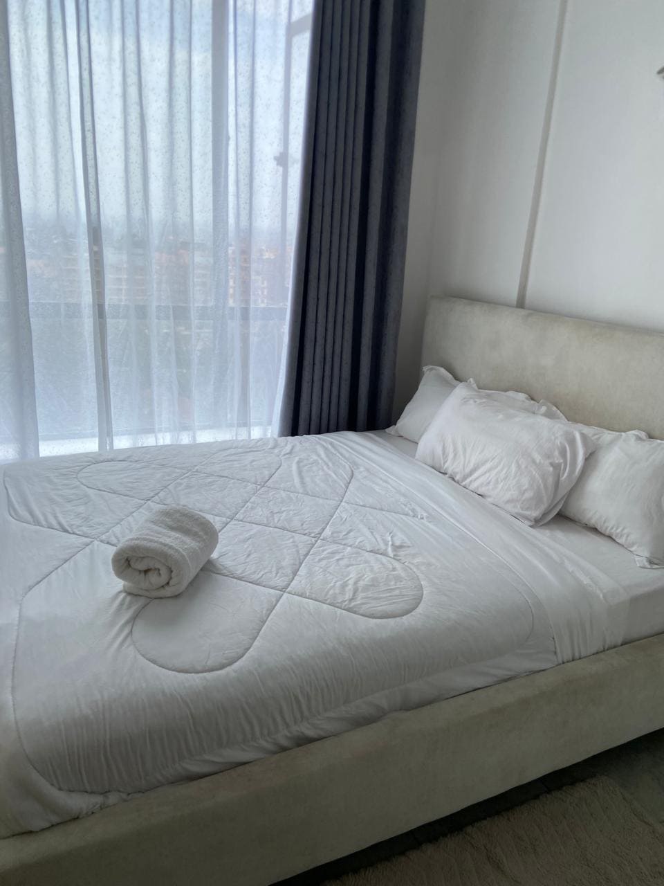 A serene bedroom features a plush queen-sized bed adorned with a soft white duvet and neatly arranged pillows. A rolled towel rests on the bed, and natural light filters through sheer curtains, enhancing the bright atmosphere of the space.