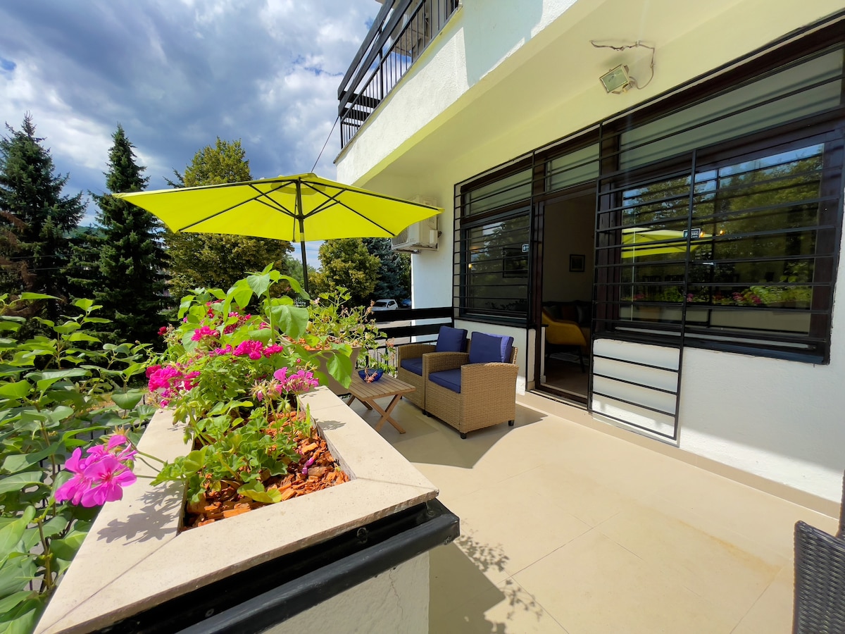 A spacious terrace showcases vibrant flower planters, with pink blooms contrasting against the natural greenery. A large umbrella provides shade over a comfortable seating area featuring two chairs and a small table, inviting relaxation amidst the scenic surroundings.