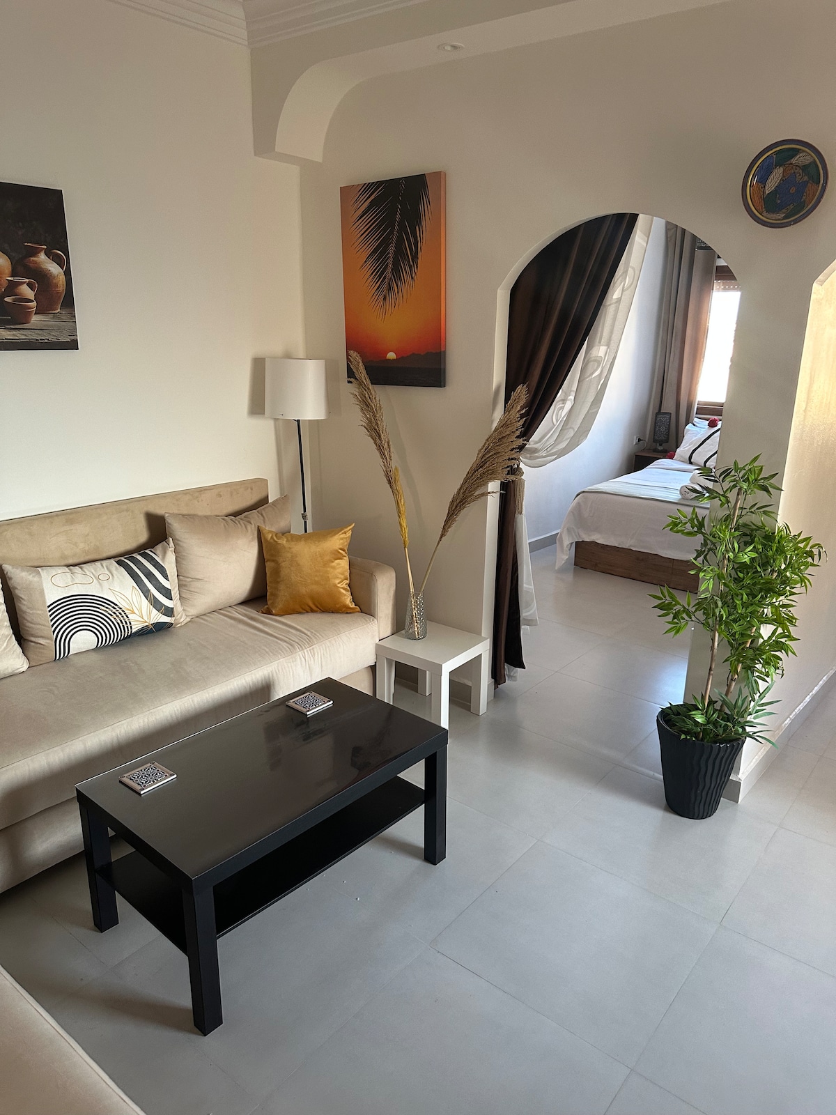 A living area showcases a light-colored sofa adorned with decorative pillows. A black coffee table sits at the center, and a tall lamp adds illumination. Wall art features a sunset and a palm, while a decorative plant brings a touch of greenery to the space.