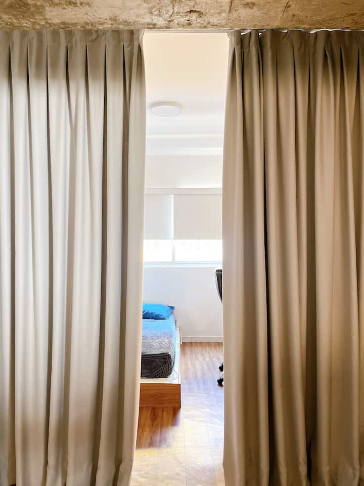 Quarto reversível separado por cortinas blackout, com cama de casal baixa e espaço de trabalho amplo 
Ambiente versátil mesa, armário e prateleiras — ideal para relaxar, trabalhar ou gravar conteúdo.