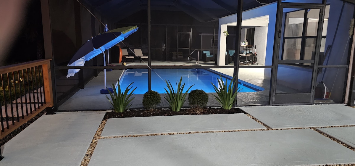A screened area showcases a peaceful private pool illuminated with soft blue lighting. An umbrella is positioned by the poolside, and sleek lounge chairs can be seen in the background. The patio features a clean path, bordered by low shrubs and decorative stones.