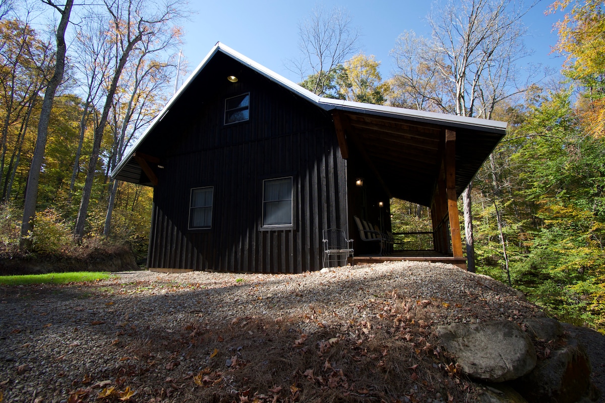 The exterior of a black cabin is surrounded by a mix of autumn foliage. Large windows reflect the natural light, while the front porch provides a shaded area with seating. The gravel pathway leads to the entrance, emphasizing the cabin's remote setting in the woods.