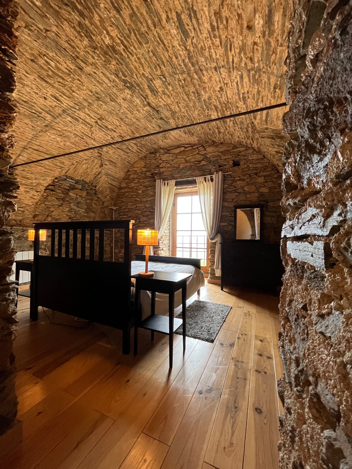 A cozy sleeping area with exposed stone walls features a spacious bed and a small table with a lamp nearby. Natural light enters through a window adorned with light curtains, while a mirror and a dresser add functional elements to the room.