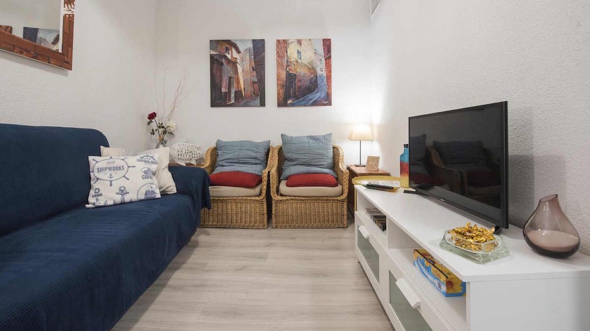 A cozy living room features a blue sofa adorned with decorative cushions. Two wicker chairs with red cushions complement the space. On the walls, two art pieces depict vibrant street scenes. A television and decorative table with snacks are present, along with soft lighting.
