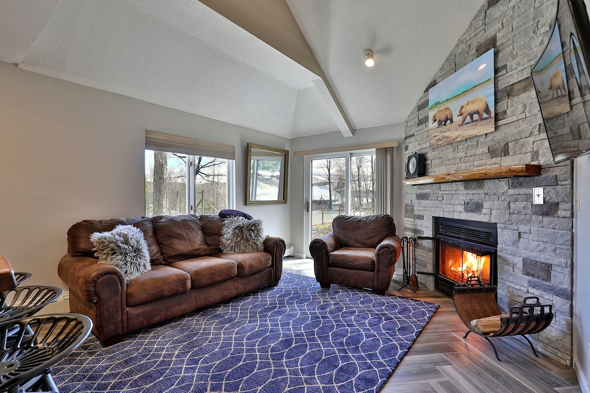 A spacious living area features a cozy sofa and armchair, set against a stone fireplace. Large windows allow natural light to fill the room, with views of the outdoor landscape. A patterned blue rug adds texture to the wooden floor beneath.