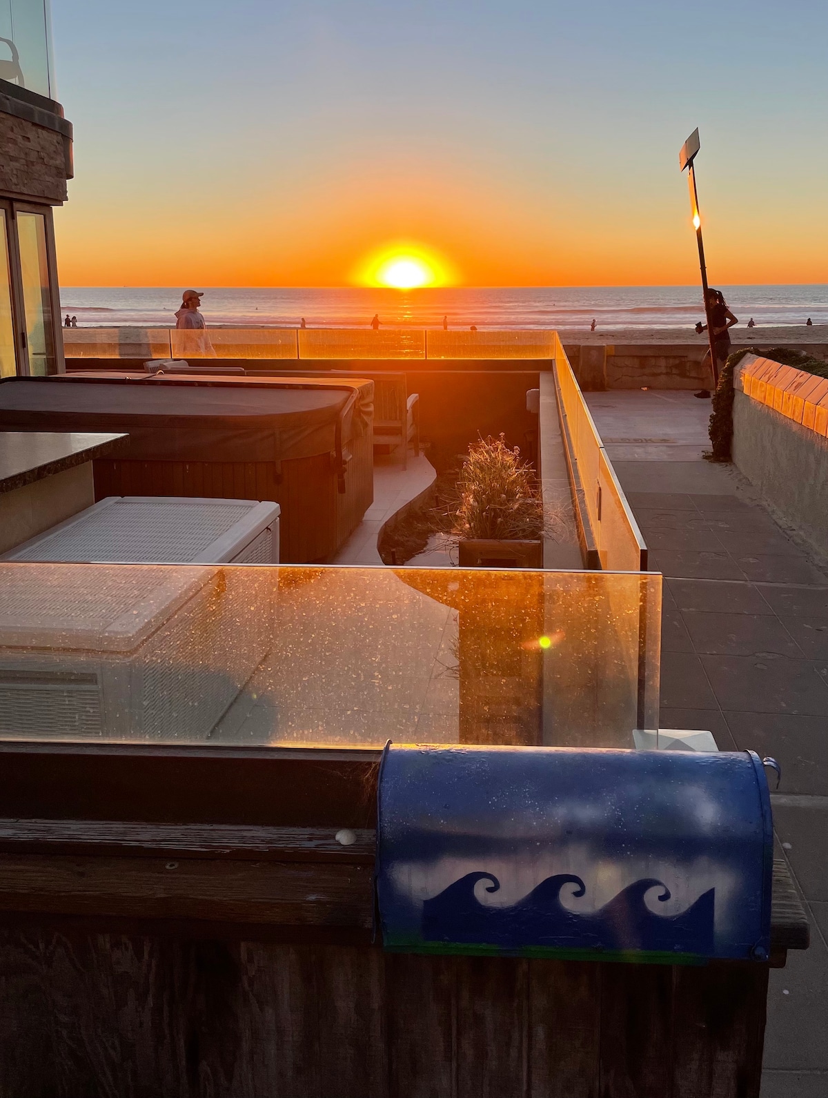 A serene sunset is observed over the ocean, casting warm orange and gold hues. In the foreground, a glass railing reflects the sunlight, while a decorative mailbox with a wave design adds a playful touch. The scene captures the tranquility of the beach atmosphere.