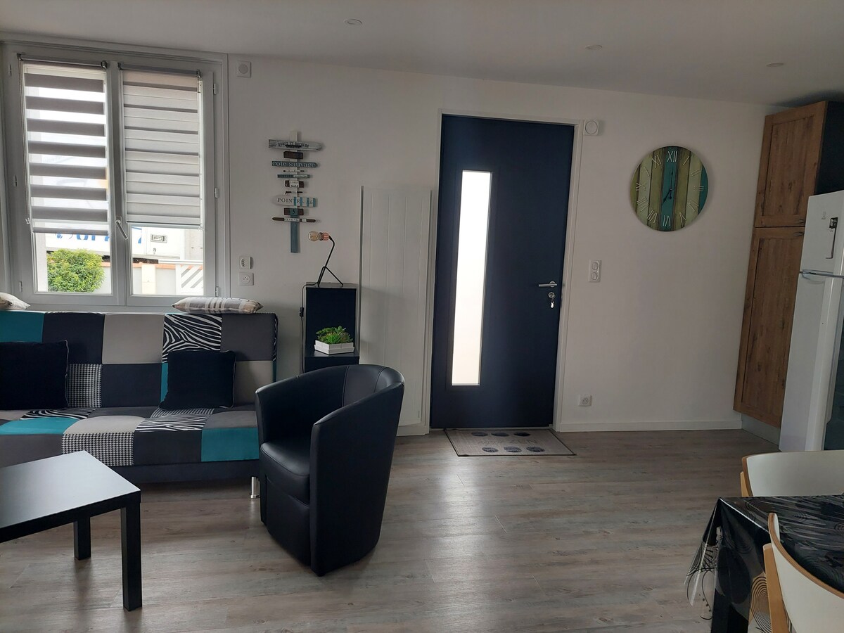 A modern living area features a comfortable patchwork couch and a round wall art piece. A sleek black chair is positioned next to a small table, and a door with a large window allows natural light to brighten the space. Light wood flooring complements the decor.