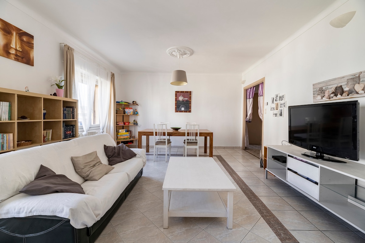 The living area features a spacious white sofa adorned with decorative pillows, paired with a simple coffee table. A dining table with white chairs is positioned nearby, while a bookshelf filled with colorful items contributes to the homey feel.