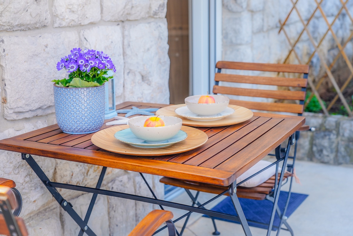 A wooden table is set on a stone patio, featuring two bowls with apples and blue plates. A potted plant with purple flowers adds a touch of color. The table is surrounded by wooden chairs, creating a welcoming outdoor dining space.