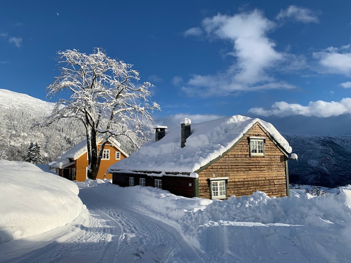 A rustic wooden house is nestled in a snowy landscape, surrounded by a thick blanket of white snow. An adjacent building displays a vibrant orange hue, contrasting with the serene blue sky and bare trees. The mountains rise gently in the background, completing the scenic view.