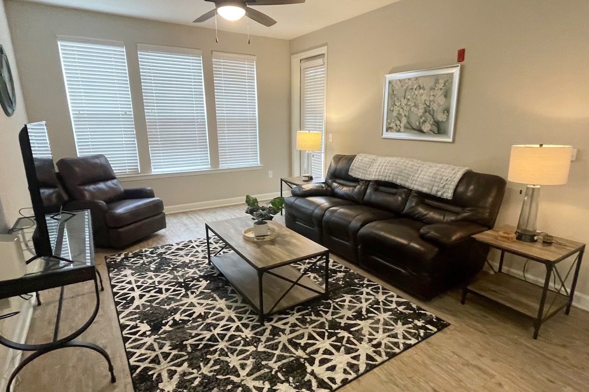 A cozy living area features a dark leather sectional sofa and a comfortable armchair. A large coffee table sits atop a patterned area rug, surrounded by natural light from the tall windows. A smart TV is positioned on a minimalist console, complemented by soft lamp lighting.
