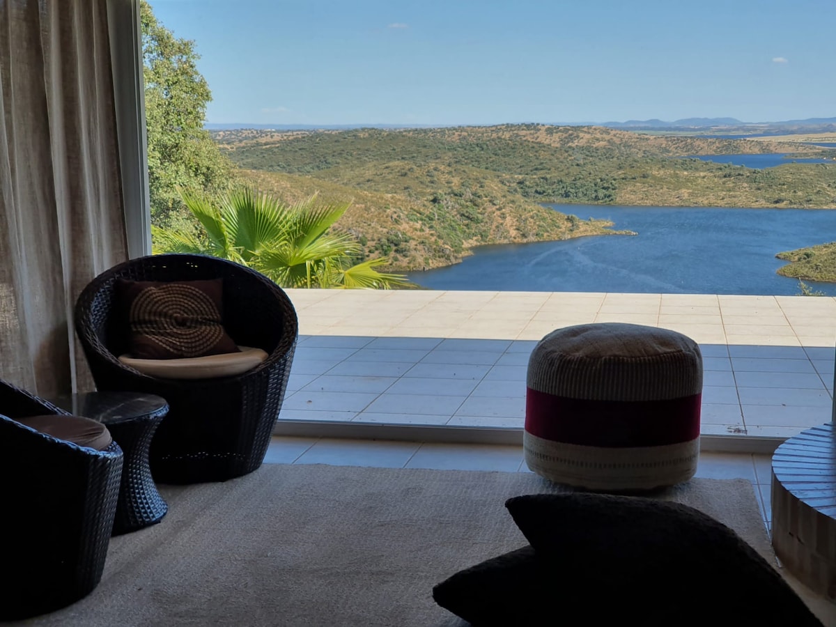 A living area features expansive glass doors that open to a stunning view of the Alqueva lake. Comfortable seating is arranged near the window, with natural light illuminating the space. Lush green hills are visible in the background, creating a serene atmosphere.