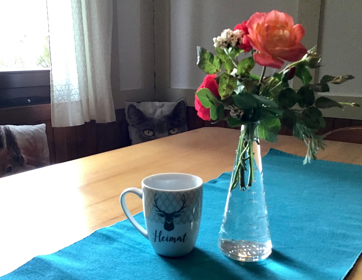 A table is set with a teal runner, featuring a clear vase filled with fresh flowers and a white mug displaying a design. Soft light filters through sheer curtains, creating a calm indoor setting.