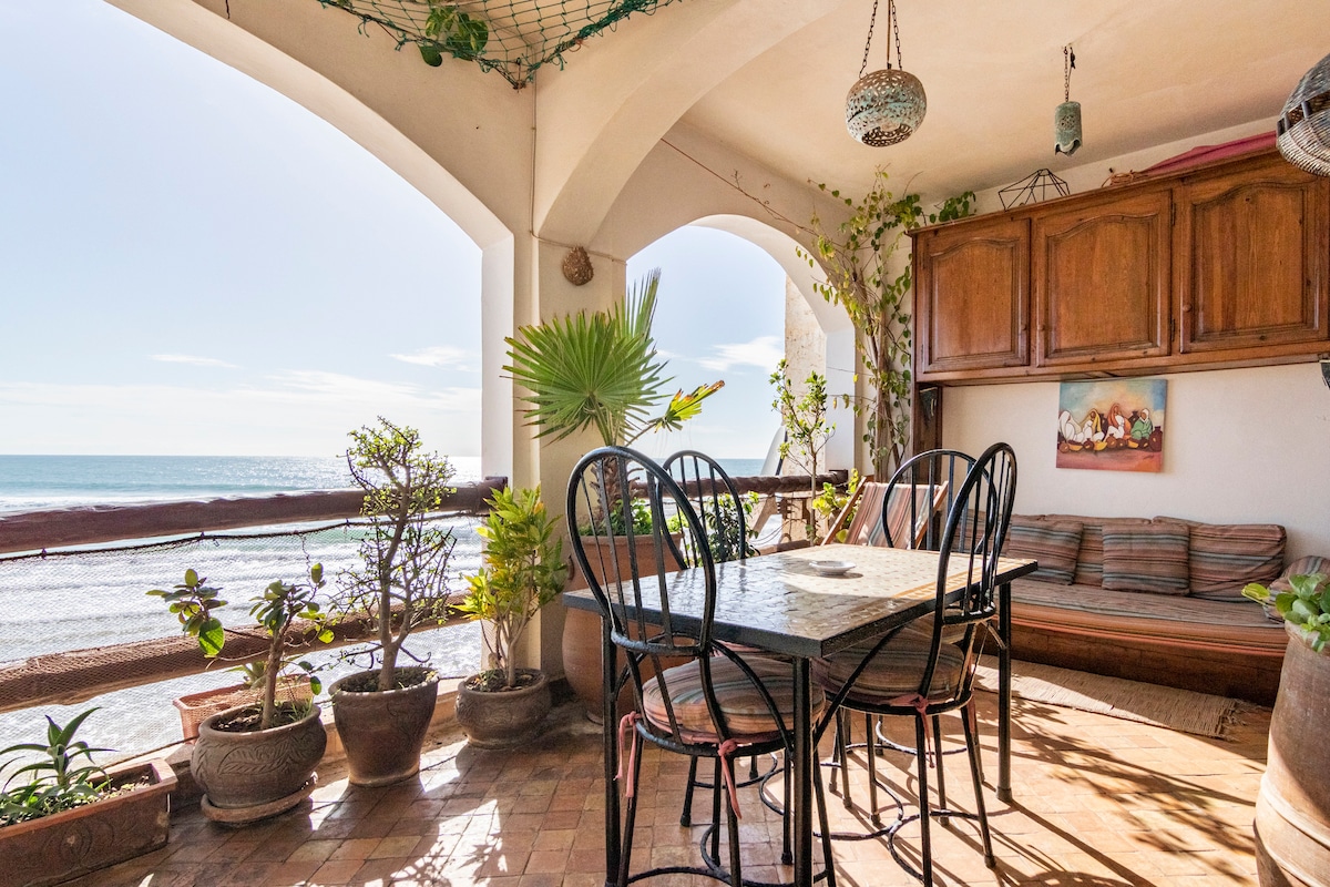 A balcony is presented with a dining table surrounded by black chairs, overlooking the ocean. Potted plants are arranged along the railing, and a comfortable seating area is visible in the corner. Natural light fills the space, enhancing the coastal atmosphere.