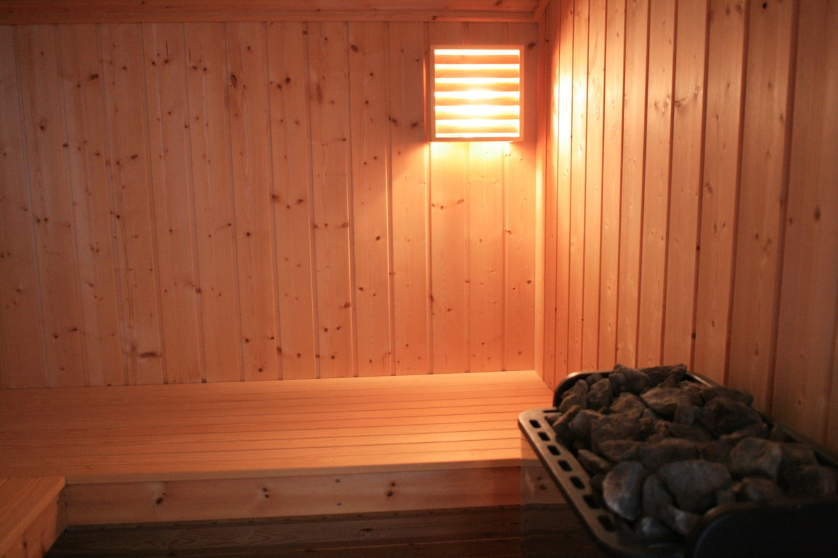 A wooden sauna interior is presented, featuring smooth cedar paneling and a heated stone stove. A bench runs along the side, providing seating. Soft light is emitted from a wall lamp, creating a serene atmosphere ideal for relaxation.