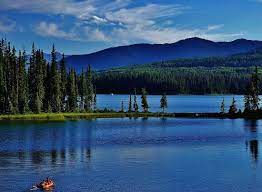 A serene lake is framed by lush green trees and rolling hills. The water reflects the vibrant blue sky, dotted with a few clouds. A canoe is visible on the water, providing a sense of calm and tranquility in the natural landscape.