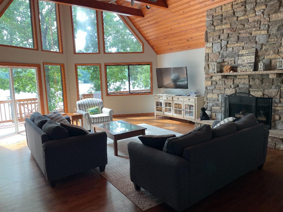 A spacious living area features large windows that provide ample natural light and views of the lake. Two gray sofas are arranged around a glass coffee table, with a cozy armchair positioned near the stone fireplace and mounted television.