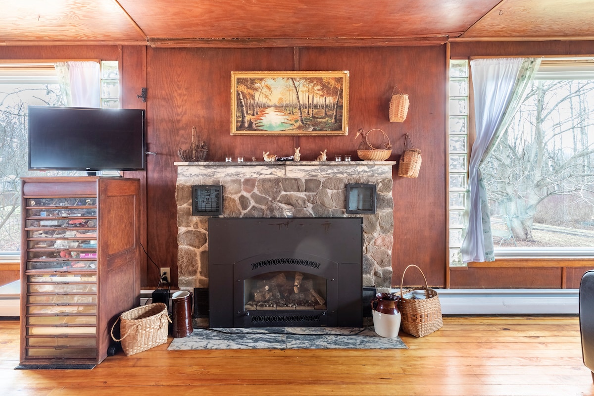 A stone fireplace is centered in the main room, framed by large picture windows. A vintage-style painting hangs above the mantel. An area with storage baskets and a television is positioned to the left, complemented by warm wooden flooring throughout the space.
