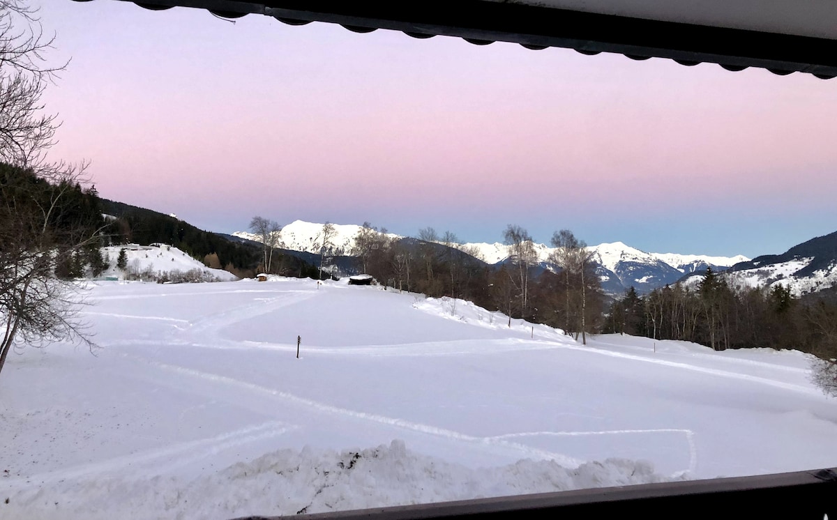 A serene winter landscape presents itself through the window, showcasing a blanket of snow covering the ground. The distant mountains are capped with snow, while a soft pastel sky transitions from pink to blue at dusk, enhancing the tranquil atmosphere.