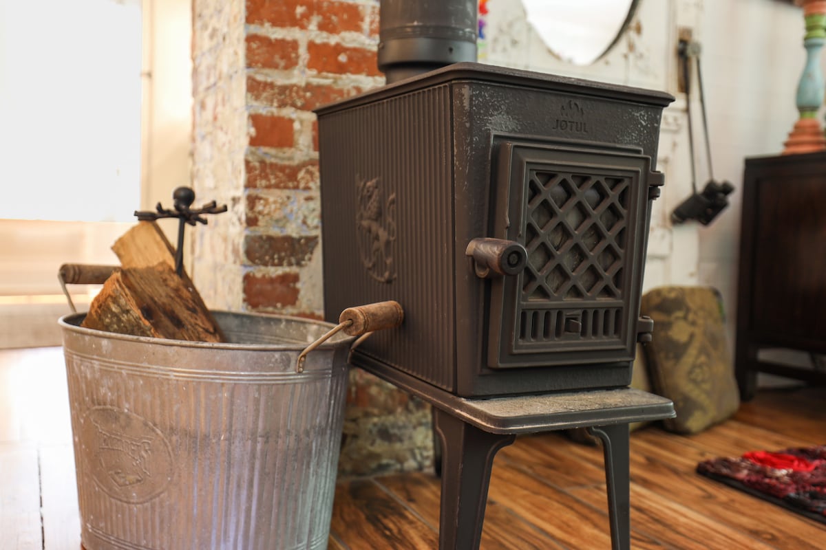 A wood stove is positioned next to a metal bucket filled with firewood. The stove features a decorative grill and is set against a backdrop of exposed brick. The warm wooden flooring highlights the cozy atmosphere of the space.