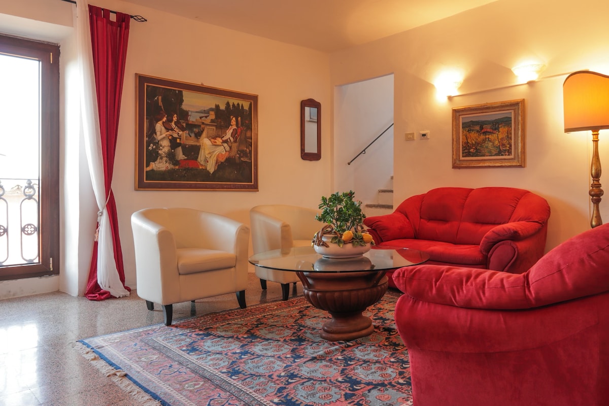 A living room features a red sofa and two white armchairs surrounding a round coffee table adorned with a decorative bowl of fruit. A traditional rug adds warmth to the floor, while a large painting hangs on the wall, complemented by natural light from a nearby window.