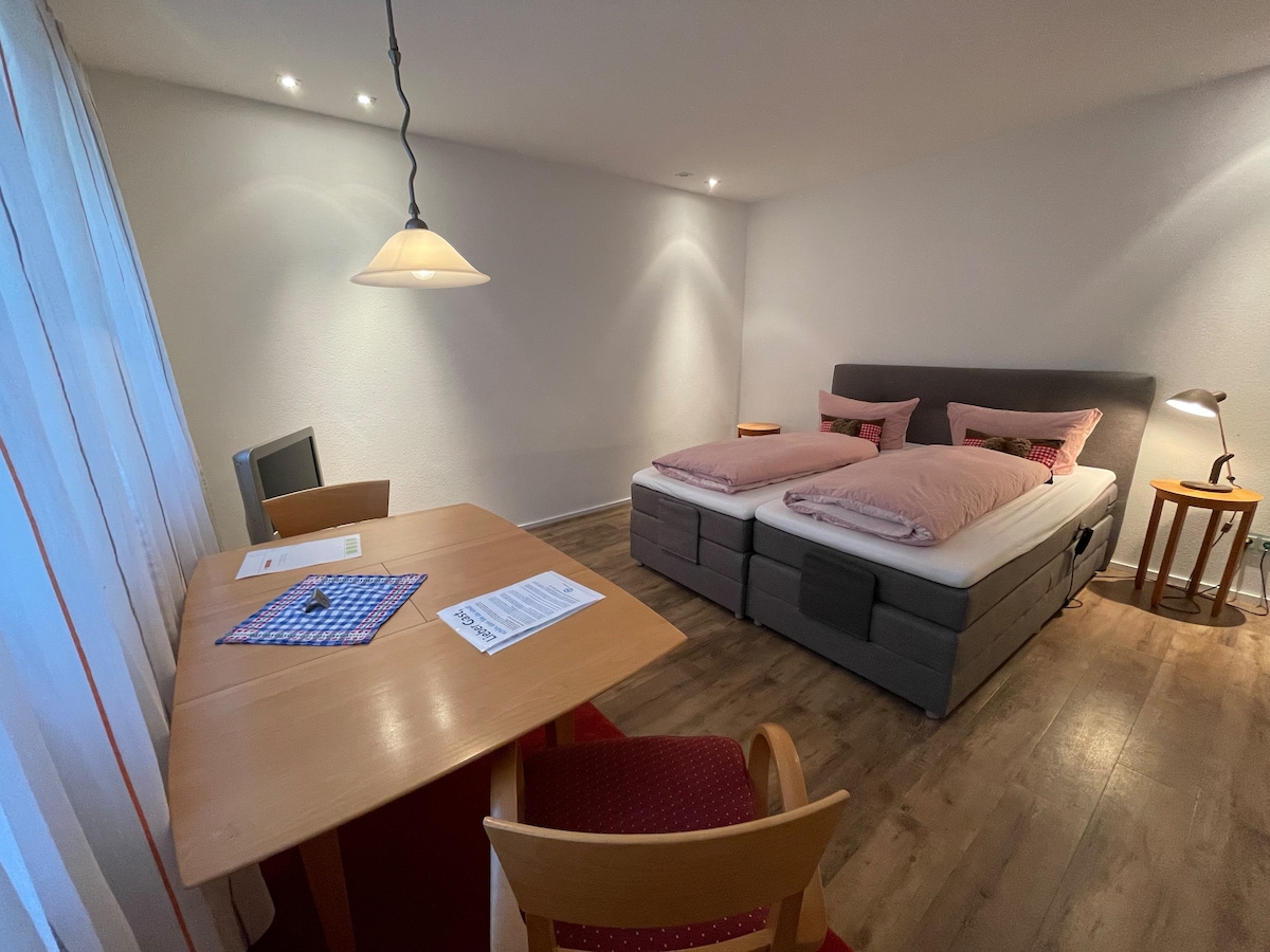 A modern sleeping area features two comfortable beds with soft linens. A wooden desk is positioned nearby, accompanied by a chair. Warm lighting from a pendant lamp and a floor lamp creates a welcoming ambiance, while sheer curtains allow natural light to filter into the space.
