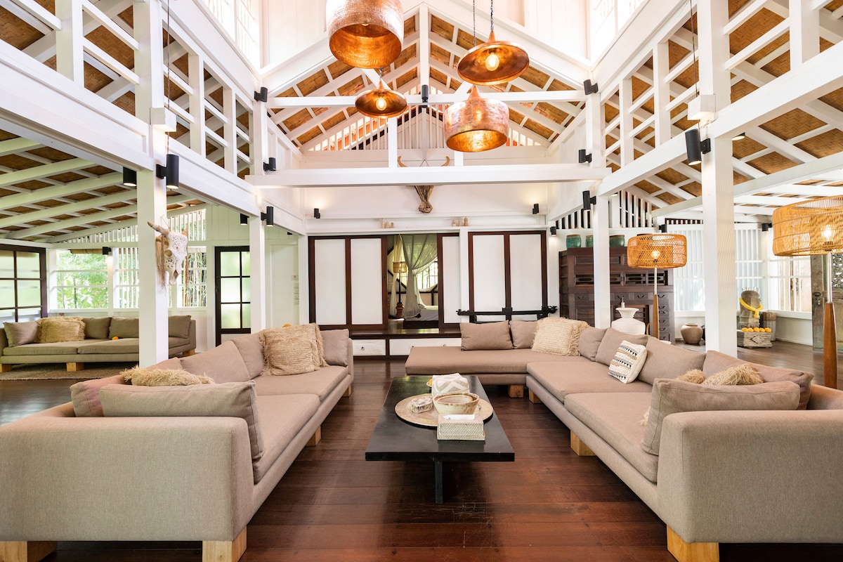 The spacious living area features a high ceiling, highlighted by decorative hanging lights. Soft beige sofas are arranged around a dark wooden coffee table, complimenting the warm wooden flooring. Large windows allow natural light to fill the room, creating an open and inviting ambiance.