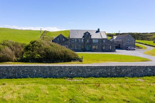 A spacious stone lodge is situated on a lush green landscape, surrounded by rolling hills and clear blue skies. The exterior features multiple windows and a slate roof, with a well-kept garden area visible in the foreground.