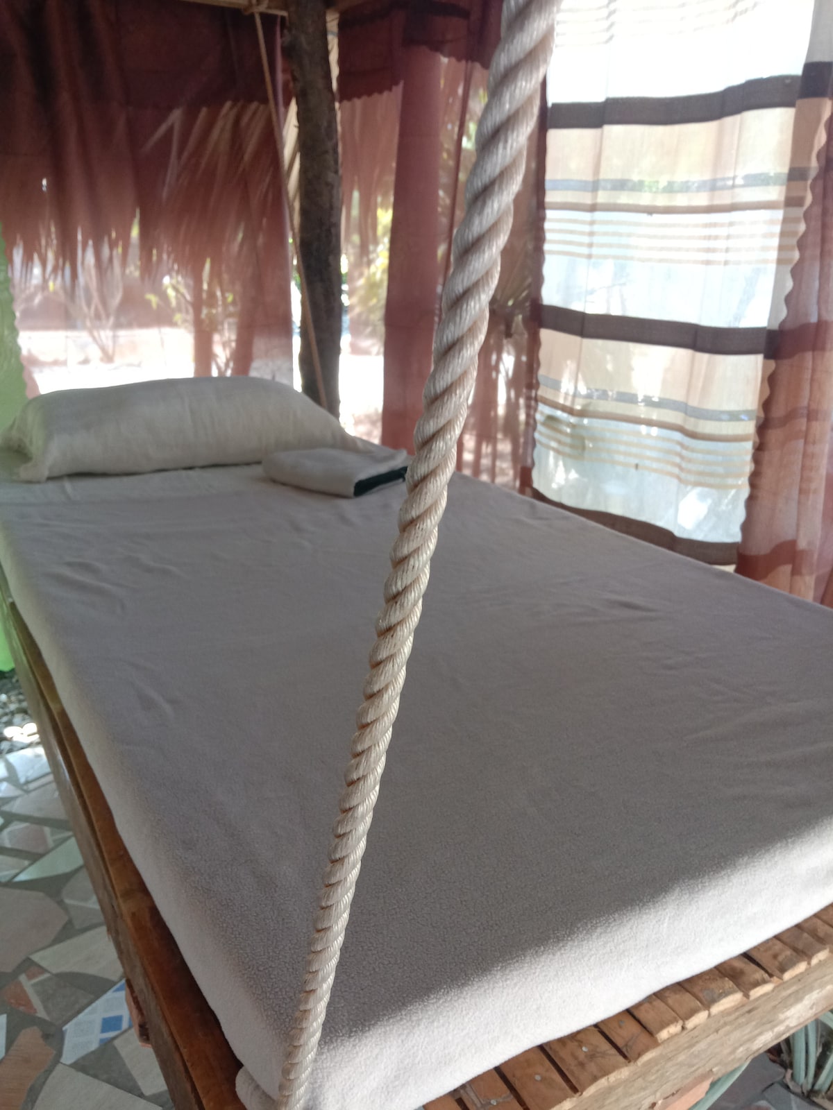 A bamboo day bed is positioned under draped fabric, allowing soft natural light to filter through. The bed features a light-colored, smooth mattress cover with a rolled towel placed beside a pillow, providing a serene place for relaxation.