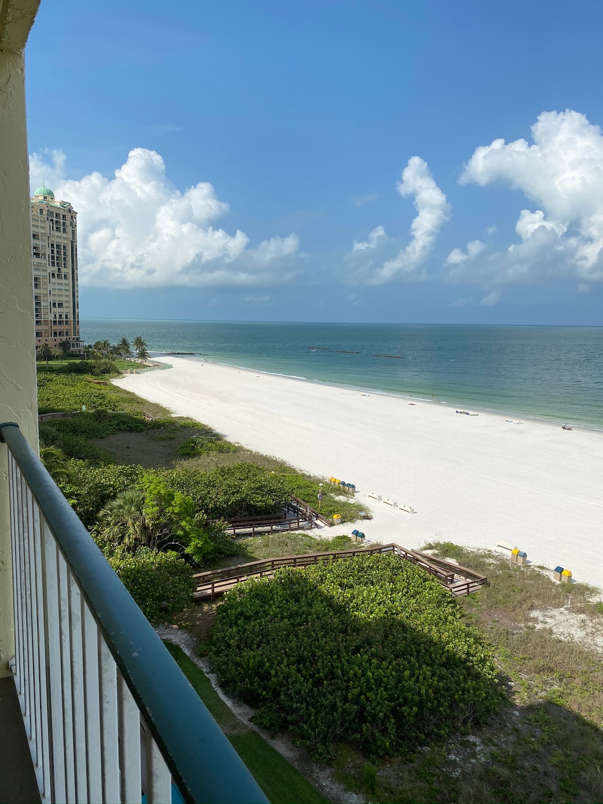 A scenic view from the balcony frames a wide stretch of pristine beach meeting the calm Gulf waters. Lush greenery borders the shoreline, while a few beach chairs can be seen in the distance. The sky displays a mix of fluffy clouds and blue hues.