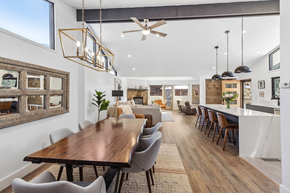A spacious dining area features a large wooden table surrounded by upholstered chairs. Natural light flows through large windows, illuminating the connected living space, which includes a cozy seating area and modern kitchen countertops. Decorative elements and a subtle color palette enhance the inviting atmosphere.