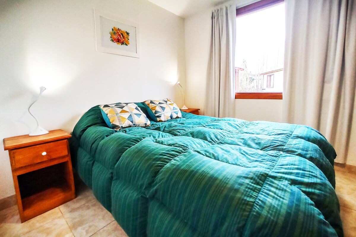 A serene bedroom features a spacious bed dressed in a green, striped comforter. Two decorative pillows rest against the headboard. On either side, wooden nightstands hold soft-lit lamps. Natural light enters through a nearby window, complemented by light curtains.