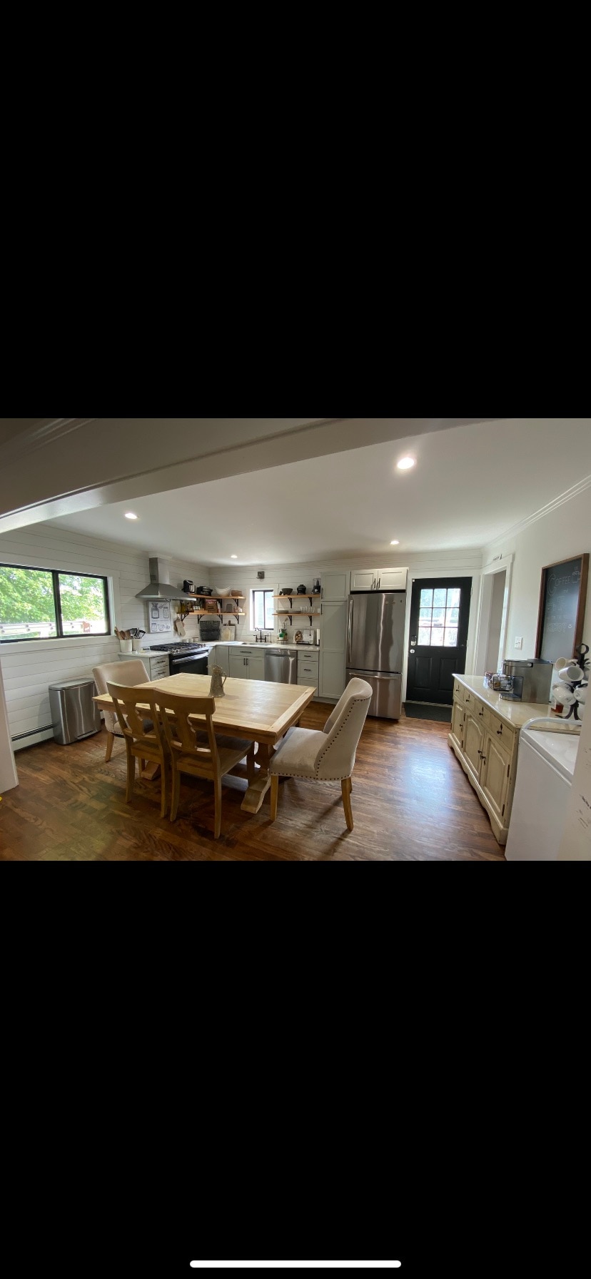 An inviting kitchen area features wooden cabinets and a spacious dining table with six chairs. Light illuminates the room from overhead fixtures. Appliances, including a gas stove and a large refrigerator, are designed for convenience. A window provides natural light, enhancing the open layout.