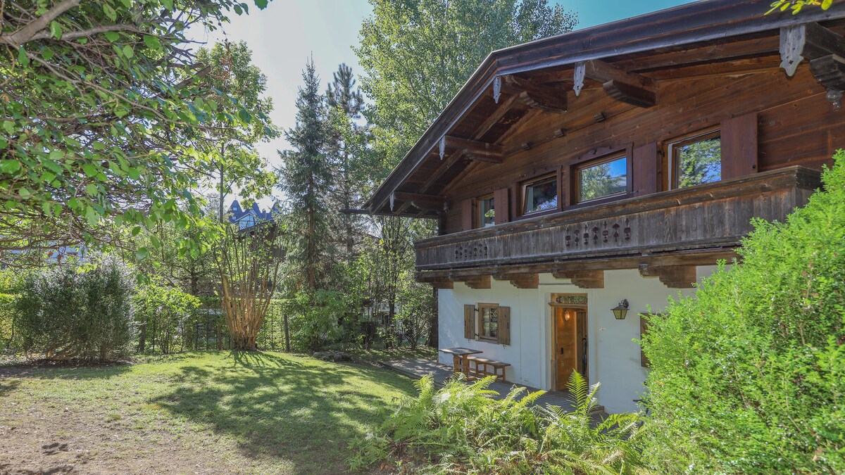 A traditional chalet is nestled among greenery, showcasing a wooden façade with a balcony. Large windows allow natural light to enter, and a small outdoor seating area is visible. Surrounding trees and shrubs provide a peaceful and secluded atmosphere.