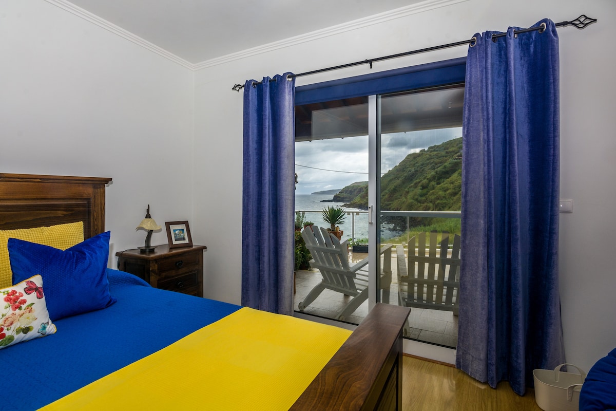 A cozy bedroom features a spacious bed adorned in bright blue and yellow linens. Large glass doors open to a serene view of the sea and coastal landscape, with sliding chairs positioned on the terrace. Natural light fills the room, accented by deep blue curtains.